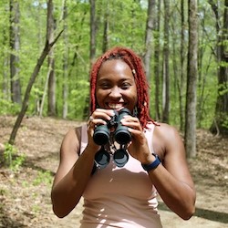 Deja Perkins smiling, holding binoculars, and facing the viewer in a wooded setting.