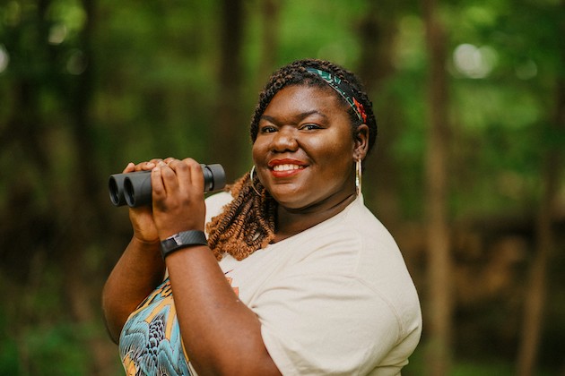 Bring Birds Back host Tenijah Hamilton smiling and holding binoculars in a wooded setting.