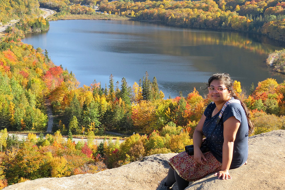Meghadeepa sits on a cliff overlooking a blue lake and forest in the background