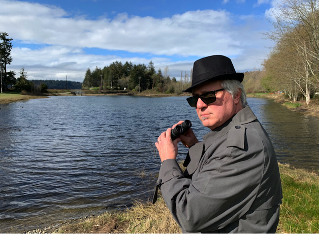 Michael Stein – Private Eye – is dressed in a trench coat, fedora, and sunglasses, and holds binoculars, looking toward the viewer. He is standing in front of a suburban pond.
