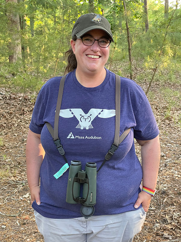 Kari Sasportas stands near a forest edge, smiling and wearing binoculars and a hat
