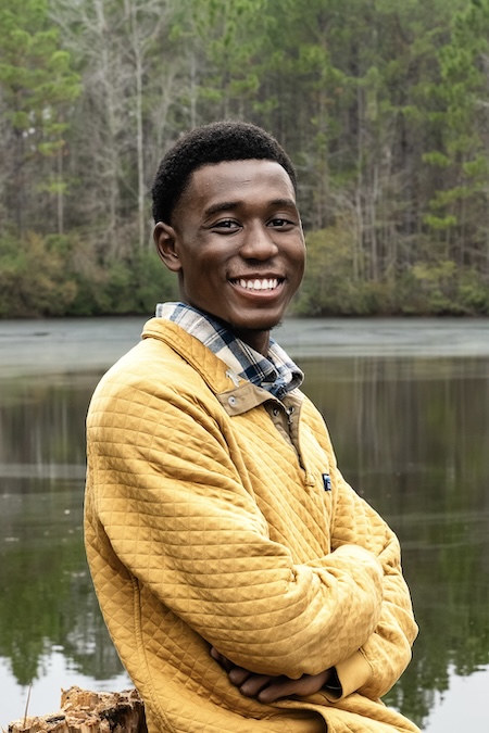 Isaiah Scott, Founder of Rookery & Roots, Scott's Wild Bird Preserve, on Bring Birds Back Isaiah Scott, a young 20-something year old, is smiling with white teeth, dressed in a yellow sweater standing in front of a lake.