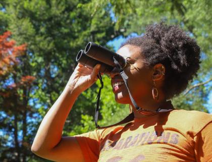 Dara Wilson in a yellow shirt looks through binoculars while standing in sunlight with trees in the background