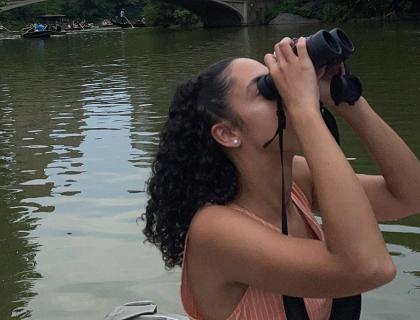 Haley Scott holding binoculars to her eyes as she looks upward while birdwatching from a boat.