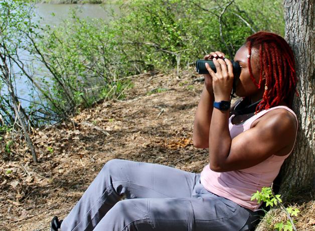 Deja Perkins sitting on the ground in filtered sunlight, with bushes and water in the background. Deja's back is against a tree trunk as she holds binoculars to her eyes, and she's wearing a pink tank top and blue pants.