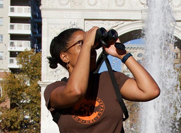 Georgia Silvera Seamans, a Black woman, raises binoculars to her face while sitting in sunlight near a fountain at the Washington Square Park in Manhattan. Georgia is the director of an organization called Washington Square Park Eco Projects.