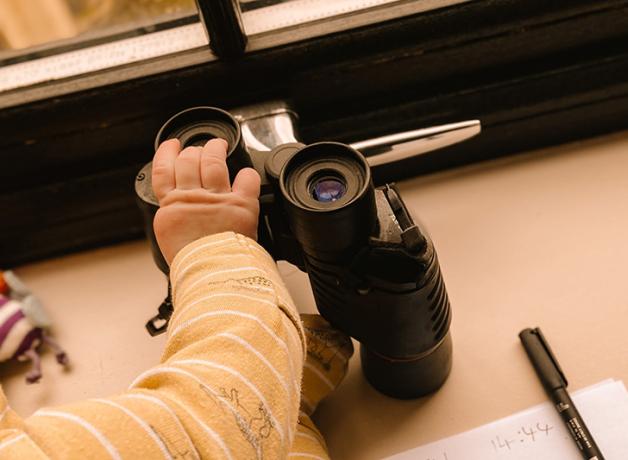 A baby's hand rests on a pair of binoculars that are on a windowsill. A pen and piece of paper are to the right of the binoculars.