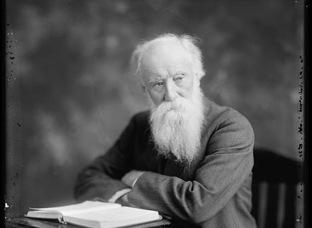 Black and white photo of American author John Burroughs, seated at a table with an open book before him