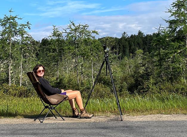 Molly Adams smiles while sitting in camping chair along a country road, her birding scope attached to a tripod. 