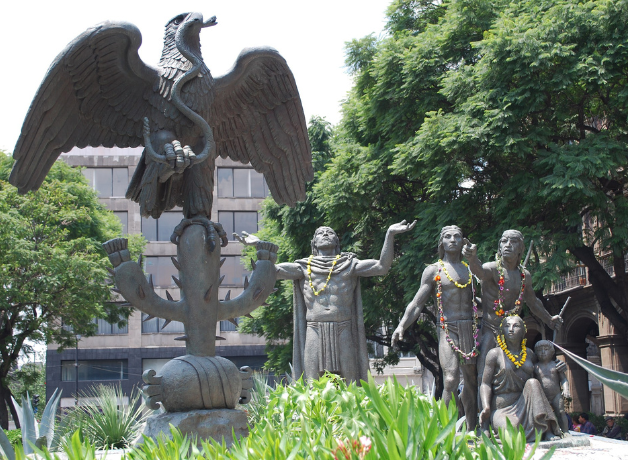 An outdoor, bronze sculpture of an eagle devouring a serpent on a cactus with a group of people looking on in awe.