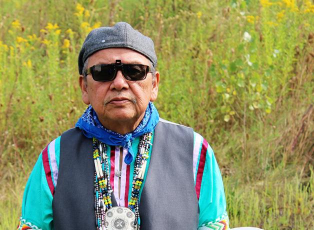 Ray Young Bear, a writer and member of the Meskwaki Nation, holding a drum and padded drumstick, with grass and wildflowers in the background.