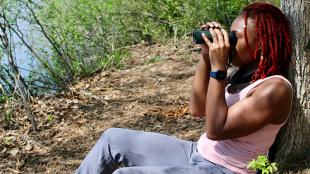 Deja Perkins sitting on the ground in filtered sunlight, with bushes and water in the background. Deja's back is against a tree trunk as she holds binoculars to her eyes, and she's wearing a pink tank top and blue pants.