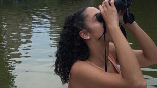Haley Scott holding binoculars to her eyes as she looks upward while birdwatching from a boat.