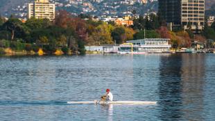 A view of Lake Merrit, with a man rowing across the water in the foreground, and trees and buildings reflecting on the water from the shore.