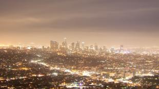 A night scene of Los Angeles seen from a nearby hill. The dense city lights make a milky glow in the sky above.