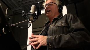 BirdNote narrator Michael Stein speaks before a microphone in a recording studio.