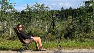 Molly Adams smiles while sitting in camping chair along a country road, her birding scope attached to a tripod. 