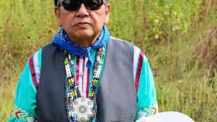 Ray Young Bear, a writer and member of the Meskwaki Nation, holding a drum and padded drumstick, with grass and wildflowers in the background.