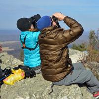 Two people sitting on rocky outcropping and looking up through binoculars on a sunny day. 