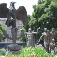 An outdoor, bronze sculpture of an eagle devouring a serpent on a cactus with a group of people looking on in awe. 