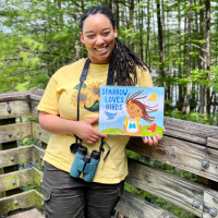A photo of Murry Burgess standing outside with binoculars and a copy of her children's book.
