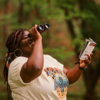 Bring Birds Back host, Tenijah Hamilton is smiling, looking up and to the right through binoculars and holding a book.